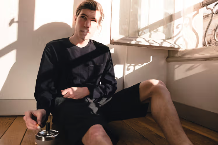 A man sitting in the sun wearing a Magenta Skateboards long-sleeve and shorts