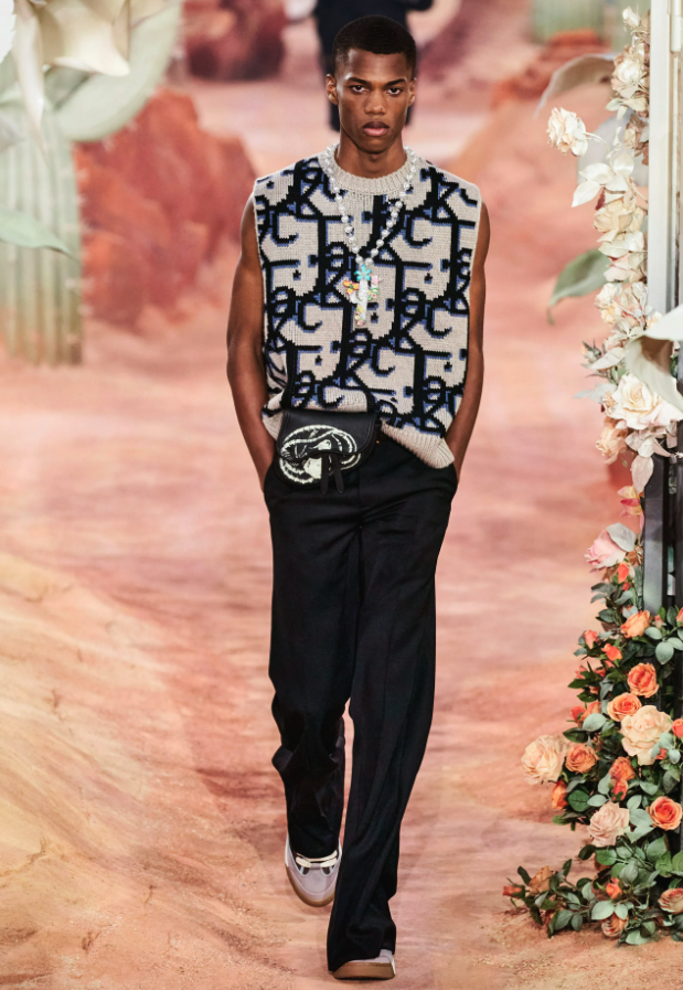A man wearing a Dior allover print vest, black pants and a colorful cross necklace