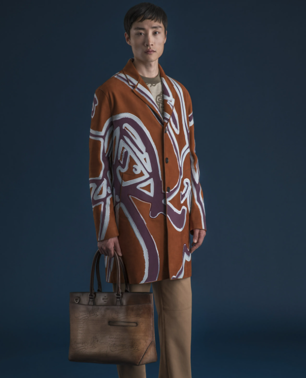 A man wearing a long Berluti jacket with a large purple pattern on top and brown pants