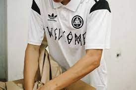 A close up of a man wearing a white Welcome Skateboards and Adidas collaboration soccer style jersey