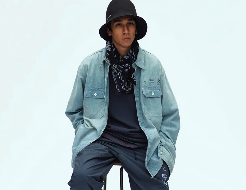 A man sitting in a Neighborhood outfit feating a navy bandana, denim overshirt, technical pants and a bucket hat
