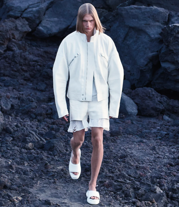 A man walking outdoors wearing an all-white Alyx jacket, shorts and slippers