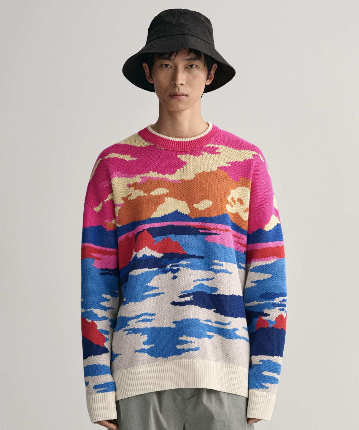 A man wearing a Gant sunset sweater, grey pants and a black bucket hat