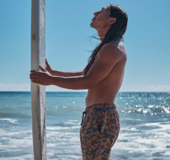 A man standing with a surf-board with colorful print swim shorts by PrAna