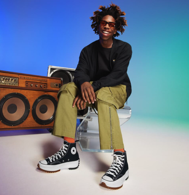 A man sitting beside a speaker wearing platform Converse in black and white