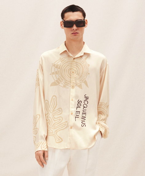 A man wearing a Jacquemus outfit featuring a cream button-up, white pants and modern sunglasses