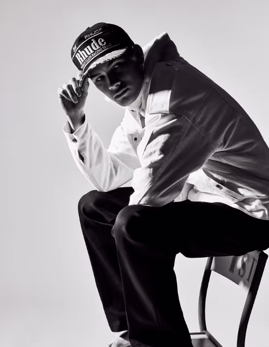A man sitting on a chair wearing a Rhude Racing hat, button up jacket and black pants