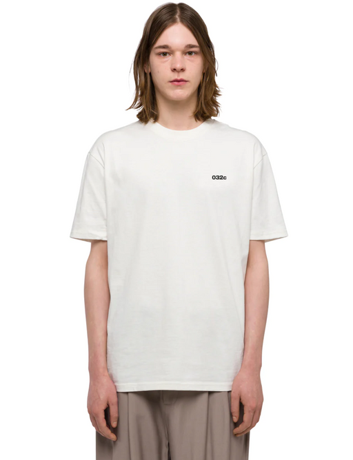A man with long-hair wearing a white 032C logo t-shirt and grey pants