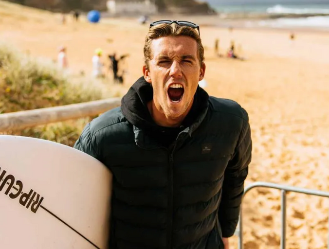 A man wearing a a Rip Curl hoodie and jacket holding a surf board