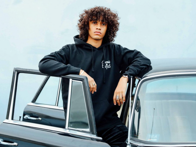A man wearing a Crooks & Castles hoodie standing beside a black car