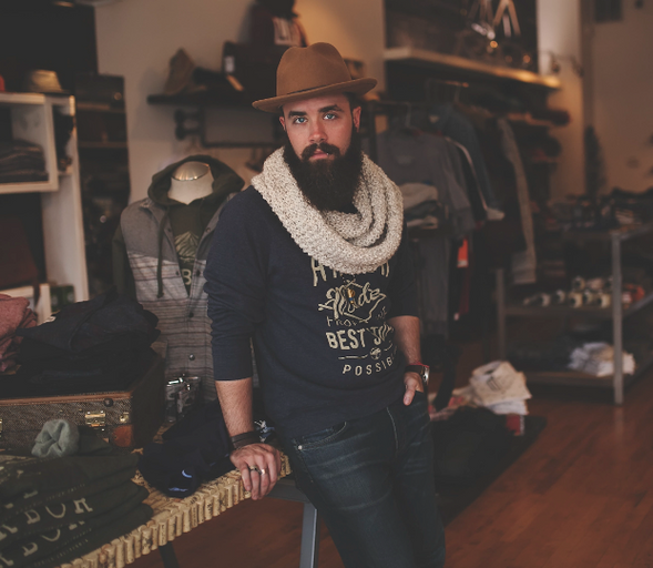 A man wearing a James Dant outfit featuring a brown hat, cream scarf and navy blue branded long-sleeve t-shirt