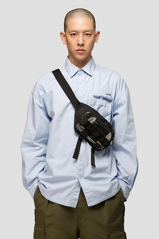 A man wearing a 4DIMENSION outfit featuring a light blue button up and a black bag slinged across chest