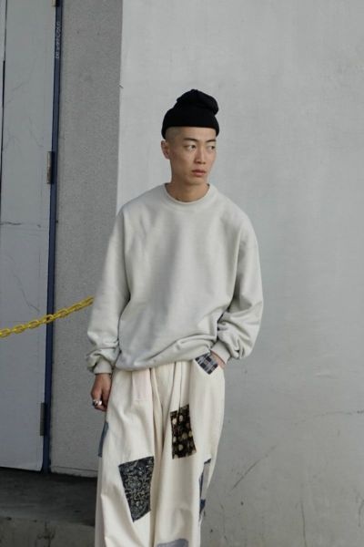 A man wearing a 1LDK grey sweater, black beanie and patched baggy pants