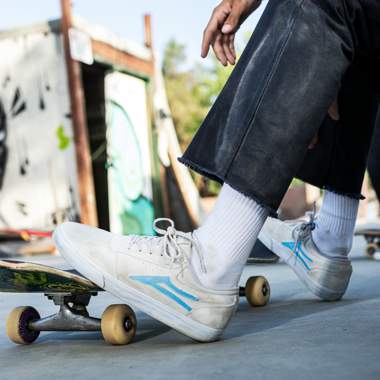 A man skateboarding wearing a cream pair of Lakai shoes with a baby blue logo
