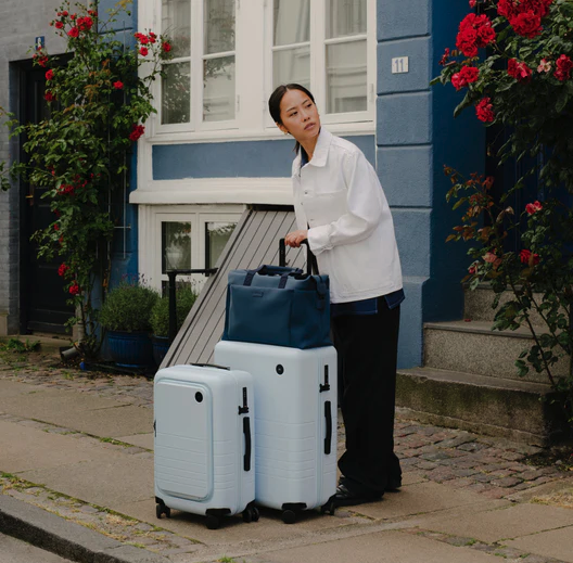 A lady holding light blue Monos roller luggage outside the front door