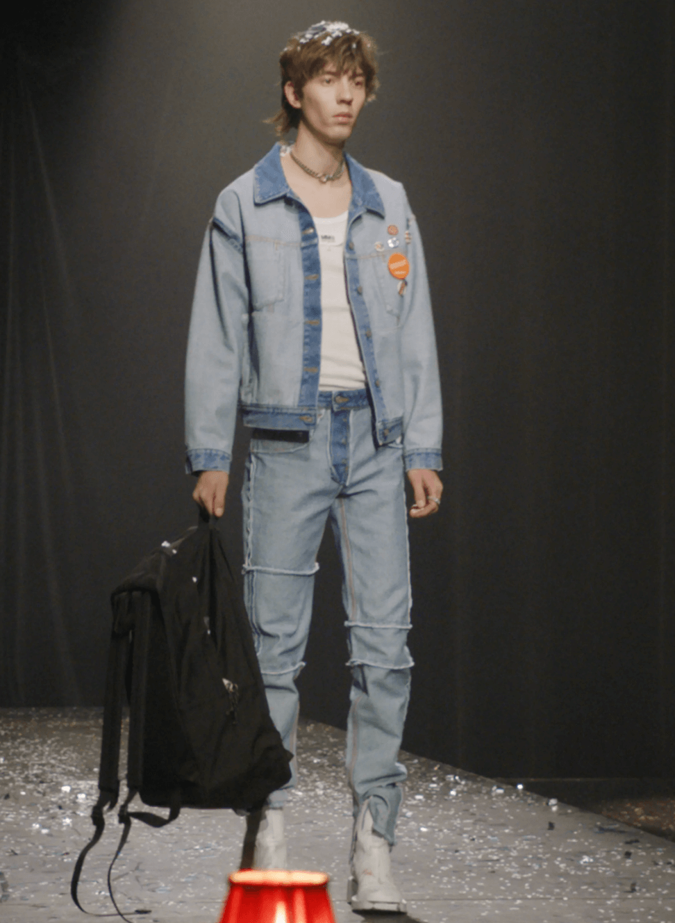 A man on a fashion runway wearing a Maison Margiela denim jacket and matching pants along with a white high-heel boots